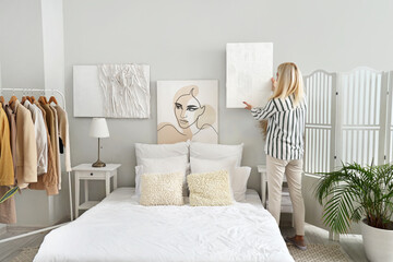 Mature woman hanging painting on light wall in bedroom, back view