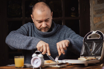 The man is having breakfast. An ordinary middle-aged man in a sweater is preparing to eat a sandwich in a dark room.