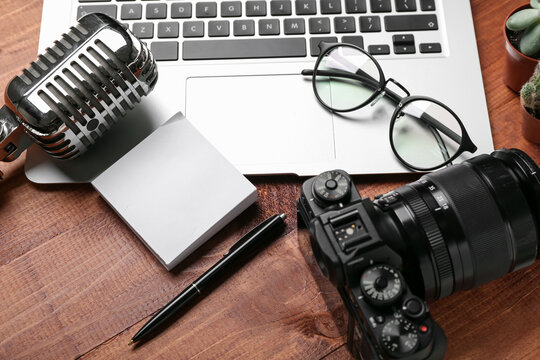 Laptop with eyeglasses, sticky notes, microphone and photo camera on wooden background, closeup