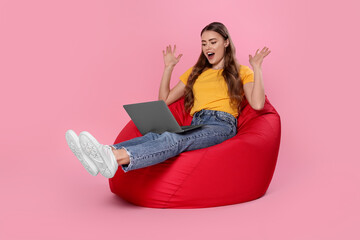 Emotional woman with laptop sitting on beanbag chair against pink background