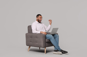 Happy young man with laptop sitting in armchair on grey background