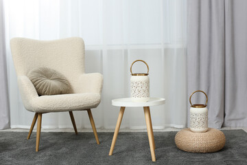 Comfortable armchair, white table and stylish lanterns near elegant curtains in light room. Interior design