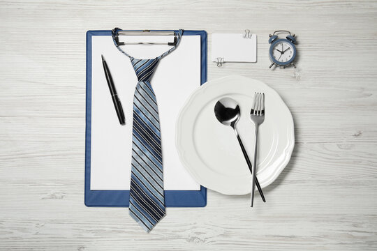Plate with cutlery, tie, clipboard and alarm clock on wooden table, flat lay. Business lunch concept