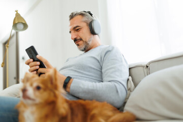 Smiling Bearded mature adult man sitting on the couch wearing headphones listening to music playing...