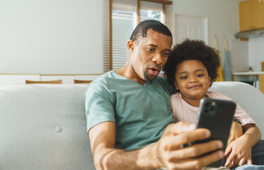 Smiling African American father and his cute boy using smartphone at home, African family doing video call on mobile phone, Black Dad and son sitting on sofa taking selfies.