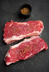 Close up of raw strip loin steaks on black cutting board with salt and spices.