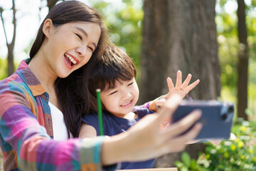 Happy cheerful Asian lovely little boy and mother sitting under the tree at park in the morning and drawing a picture and taking a selfie photography on smartphone together.