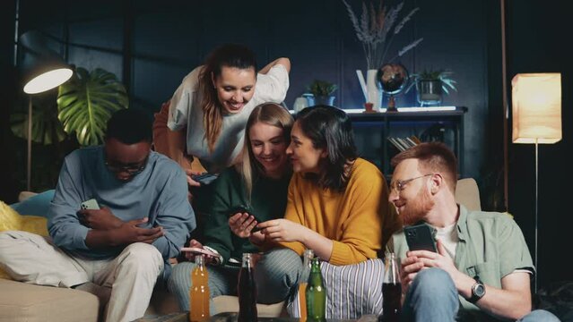 Young beautiful woman shows funny meme to multiethnic friends using smartphones on couch at home. Fun together.