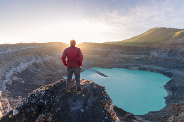 Mountaineer man looking at the Poas Volcano Crater Lagoon at sunrise surrounded by volcanic rocks...