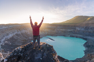 victorious mountaineer man in front of Poas Volcano Crater Lagoon at sunrise surrounded by volcanic rocks in Poas Volcano National Park in Alajuela province of Costa Rica - obrazy, fototapety, plakaty