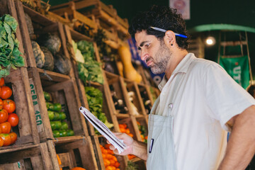 Middle-aged Latin man with a pen in his ear and a list in his hand puzzled because the inventory does not match the products on the shelf.