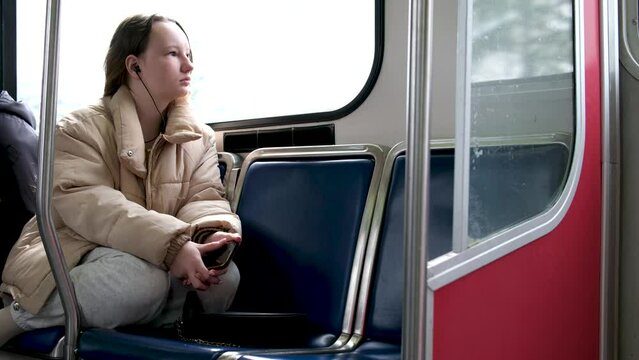 teenage girl rides in skytrain on train in bus headphones in ears listens to music autumn spring winter warm beige jacket hair is tied down real person sits tucked legs under sad dreams awaits stop