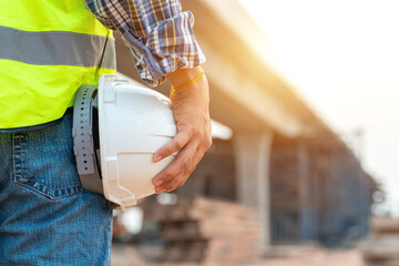 Close-up helmet of Construction worker or civil engineer holding safety white helmet and construction drawing against the background of road construction site.