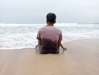 A man sits on the edge of a white sandy beach and looks out to sea.