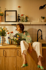 Cute and relaxed woman sits on kitchen sink in modern kitchen interior decorated with fresh...