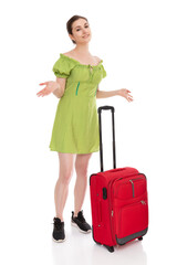 Full length portrait of perplexed girl in green dress with red suitcase to travel, isolated on white background. Young woman waiting for transport and spreads her arms to the sides, posing in studio.