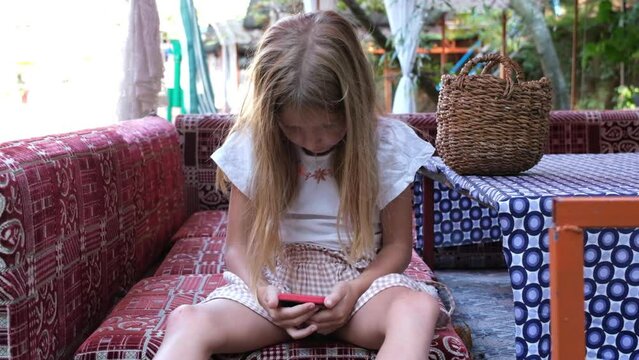 Cute little girl sits in an unhealthy position and uses a mobile phone. Harm to health from electronic device.