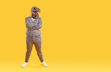 Funny eccentric overweight man posing in dinosaur mask on his head and in pajamas with leopard print. Bizarre fat man stands with crossed near copy space on orange background. Full length. Banner.