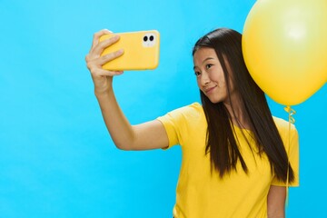 Asian woman holding the phone and looking at the screen talking on a video call with a yellow balloon in her hands on a blue background in a yellow T-shirt smiling with teeth 