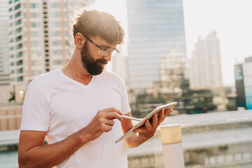 Outdoor portrait of Handsom  man in white t-shirt using  tablet , searching in internet, standing over moden city background with  skyscraper.