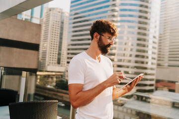 Handsome man in white t-shirt using  tablet , searching in internet, standing over moden city background with  skyscraper.