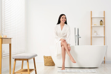 Beautiful happy woman in stylish bathrobe sitting on tub in bathroom - obrazy, fototapety, plakaty
