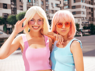 Two young beautiful smiling hipster female in trendy summer clothes. Sexy carefree women posing in the street in wigs. Positive funny models having fun at sunset. In sunglasses. At sunny day