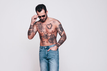 Portrait of handsome confident stylish hipster lambersexual model in jeans. Sexy modern man. Naked torso with tattoos. Fashion male posing in studio. Isolated on white. In sunglasses
