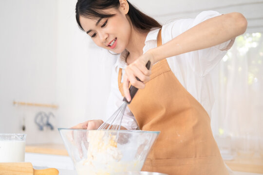 Young fun housewife housekeeper chef baker woman in light brown apron work at table kitchenware use whisk mixing flour baking cake in kitchen .Process cooking food concept Woman baking at home. - Powered by Adobe