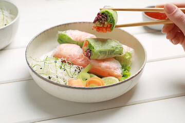 Woman eating delicious spring rolls wrapped in rice paper with chopsticks at white wooden table, closeup