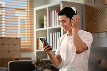 Fototapeta na wymiar Happy Asian man enjoy favorite songs in headphones and using smartphone on couch. Technology and lifestyle concept