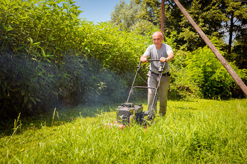 Farmer is mowing lawn in the garden with a petrol lawn mower that smokes