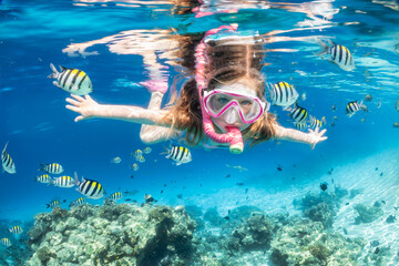 A little girl with mask and snorkel enjoys the underwater life of the tropical ocean wth colorful...