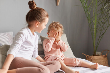 Charming elder sister taking care of her younger infant baby happy little girls playing hide and seek in bedroom cute kid with hair bun helping mother to play with baby child.
