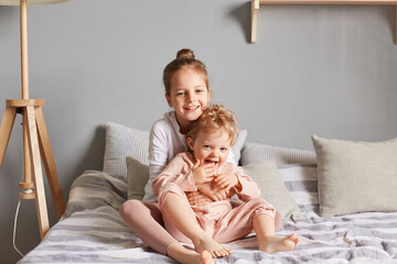 Positive adorable cute happy little girls playing in bedroom, sitting on bed hugging and smiling, elder sister taking care of toddler baby during weekend.