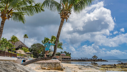 Obraz premium A palm tree leaned over the beach. A man in sunglasses and a cap is standing on a curved trunk. In the distance - the buildings of the hotel, the ocean at low tide. Clouds in the sky. Seychelles. Mahe