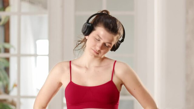 A woman in red sportswear puts on headphones and starts her morning workout. A young brunette listens to music on headphones during a morning workout. The woman kneads her shoulders and neck.