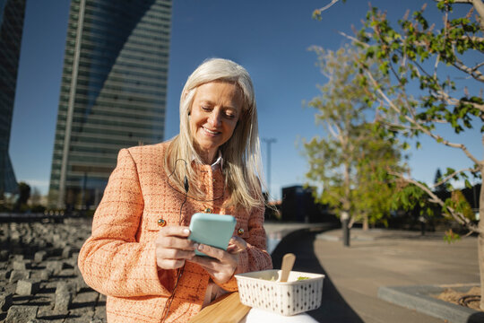 Smiling businesswoman using smart phone sitting on bench at sunny day