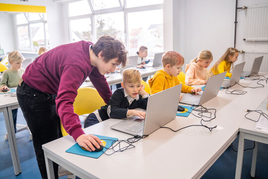 Volunteer assisting boy using laptop sitting at desk with friends in school