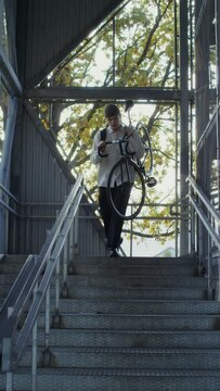 A young man of European appearance in a knitted hat, with a backpack on his back carries a bicycle down a metal ladder, hanged it on his shoulder