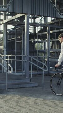 A young man of European appearance in a knitted hat with a backpack on his back rides a bicycle to a metal ladder, dismounts, takes a bicycle in his arms and carries it up the steps