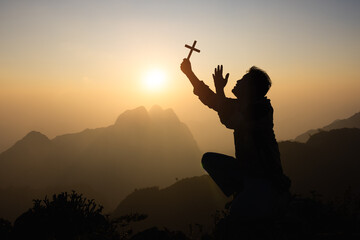 Silhouette of faithful man praying with Christian cross at sunset as concept for religion, faith,...