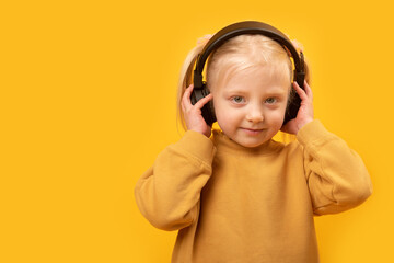 Young fair-haired girl with headphones on yellow background. Child enjoys listens to music in headphones.