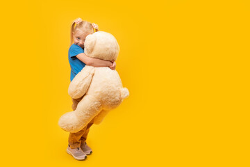 Little blond girl with tails hugs large toy bear. Studio portrait of child with toy on bright...