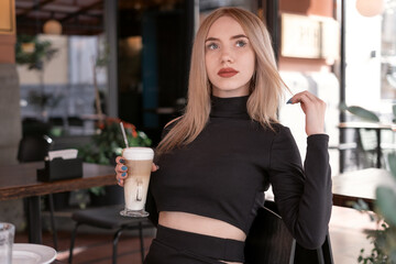 Brooding attractive blonde woman sits in coffee shop with cup of cappuccino coffee latte. Beautiful girl alone in cafe