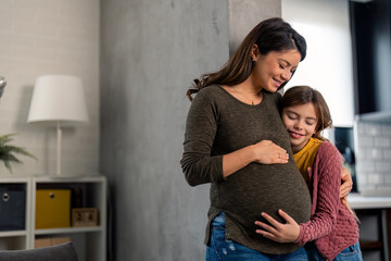 Shot of a young girl cuddling her pregnant mother’s stomach while standing together at home. Mom bonding with child in the living room.