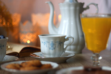 Cup of tea or coffee, plate of cookies, cup of blueberries, plate of chocolate, glass of juice, book, reading glasses, teapot, flowers and lit candles on the table. Brunch or afternoon tea concept.