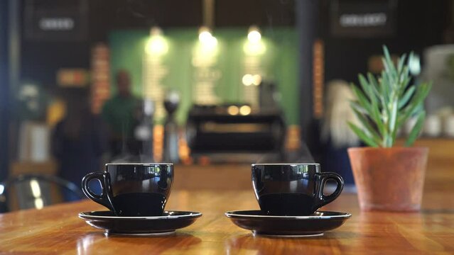 A shot of two steaming cups of hot coffee being served. The cups are in focus as the hands serving move out of the frame. In the background, a Barista works behind a coffee machine, out of focus.