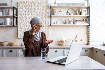 A sick woman in a hijab in the kitchen drinks medical pills and drinks water, a Muslim woman works remotely at home and uses a laptop.