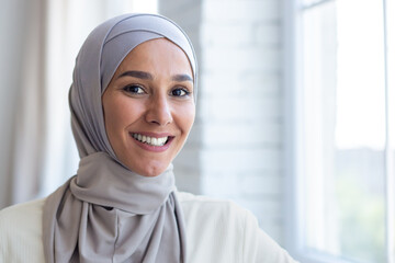 Close-up portrait of young beautiful arab woman, muslim woman in hijab smiling and looking at camera, standing near window at home.